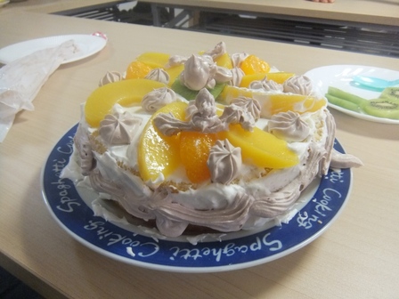表面をクリームで覆った丸いスポンジケーキの上に缶詰のみかん・黄桃と輪切りのキウィフルーツを載せ、クリームでデコレーションしたケーキ。