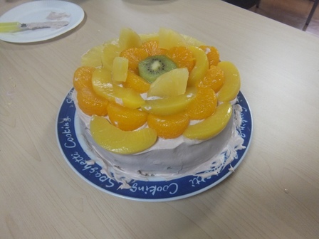 表面をクリームで覆った丸いスポンジケーキの上に缶詰のみかん・パイナップル・黄桃をこぼれそうなほど載せ、中央に輪切りのキウィフルーツを1個載せたケーキ。