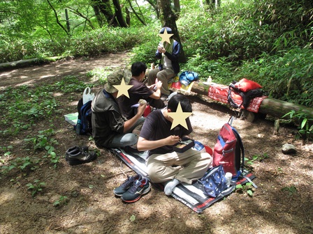 4人の参加者が、木陰の地面と丸太に敷いたシートに座って昼食をとっている。